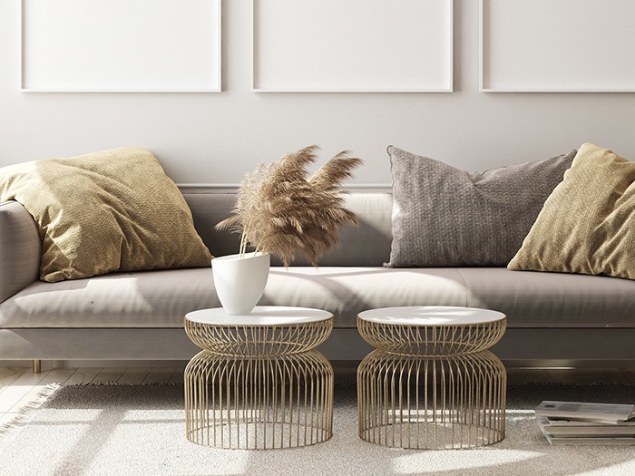 Gray velvet sofa with large fashionable cushions in beige and gray with two gold tables with marble tops and a a white vase holding pampas grass. 